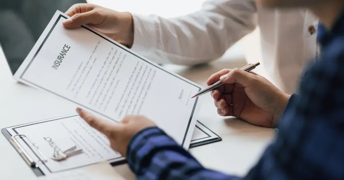 A person in a blue checkered shirt holds a document titled "Insurance" while another person, partially visible, is handing over a similar document. Both documents are on a clipboard on a table.