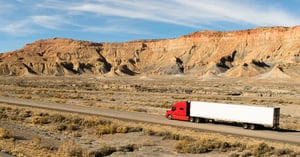 Red semi-truck with a white trailer driving along a desert highway with rocky cliffs and mesas in the background.